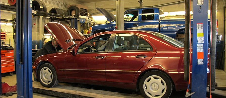 A technician under the raised hood of a red Mercedes on a lift in the Ness Auto Service bay